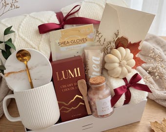 May include: A gift set containing a white ribbed mug with a marble coaster and gold spoon, a box of Lumi creamy hot chocolate, a tube of shea cream, a jar of bath salts with a wooden scoop, a pumpkin-shaped candle, and a cream-coloured knit blanket.