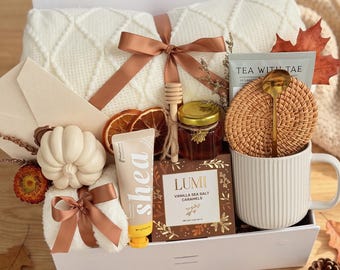 May include: A gift box containing a white cable-knit blanket tied with a brown ribbon, a white ceramic mug, a gold spoon, a rattan coaster, a jar of honey with a wooden dipper, a shea butter tube, a box of Lumi vanilla sea salt caramels, and a pumpkin.