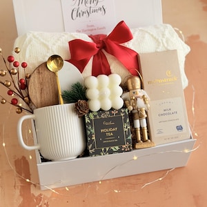 May include: A white gift box containing a cream blanket, red bow, mug, candle, tea, chocolate, and a gold nutcracker ornament. A card reads "Merry Christmas" and "From The Harringon Family."