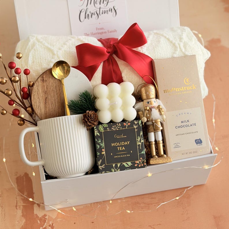 May include: A white gift box containing a cream blanket, red bow, mug, candle, tea, chocolate, and a gold nutcracker ornament. A card reads "Merry Christmas" and "From The Harringon Family."