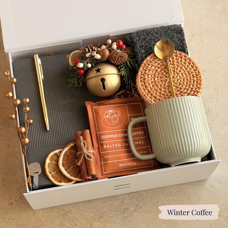 May include: A white gift box containing coffee-related items. It features a green mug, coffee sachet, cinnamon sticks, dried orange slices, a gold spoon, a woven coaster, a gold pen, and a decorative bell. The text "Winter Coffee" is at the bottom.