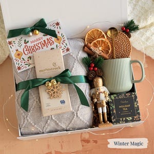 May include: A festive gift box containing a grey blanket, a green mug, and a Christmas card. The box also includes a nutcracker, tea, and decorative items. It's tied with a green ribbon and a snowflake ornament, perfect for the holiday season.