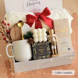 May include: A white gift box containing a mug, tea, chocolate, candle, and a decorative nutcracker. A red bow sits atop a cream-coloured blanket. The box includes a gold spoon, a wooden coaster, and berries. The text "Merry Christmas" is visible.