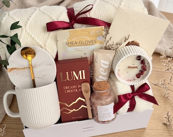 May include: A gift set containing a white ribbed mug with a marble coaster and gold spoon, a box of Lumi creamy hot chocolate, a wooden scoop, a jar of bath salts, a shea butter glove packet, a candle with dried petals, and a white blanket.