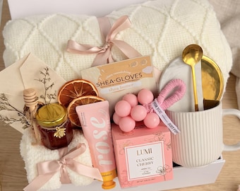 May include: A gift box containing a cream-coloured knit blanket, a jar of honey with a wooden dipper, two dried orange slices, shea butter gloves, a rose hand cream, a pink beaded accessory, a box of Lumi Classic Cherry tea, and a white ribbed mug.