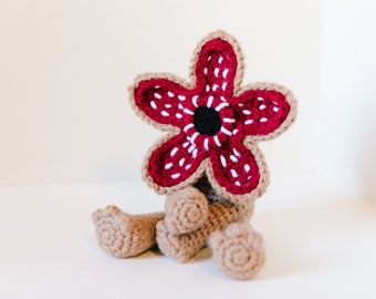May include: A crocheted plush toy shaped like a creature from the show Stranger Things. The creature has a red and white flower-like head with a black center.
