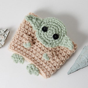 May include: A handmade crochet cup cozy featuring a green and tan design resembling a character from a popular science fiction franchise. The cozy has black button eyes and green accents. Toy models of a spaceship and a star destroyer are also present.