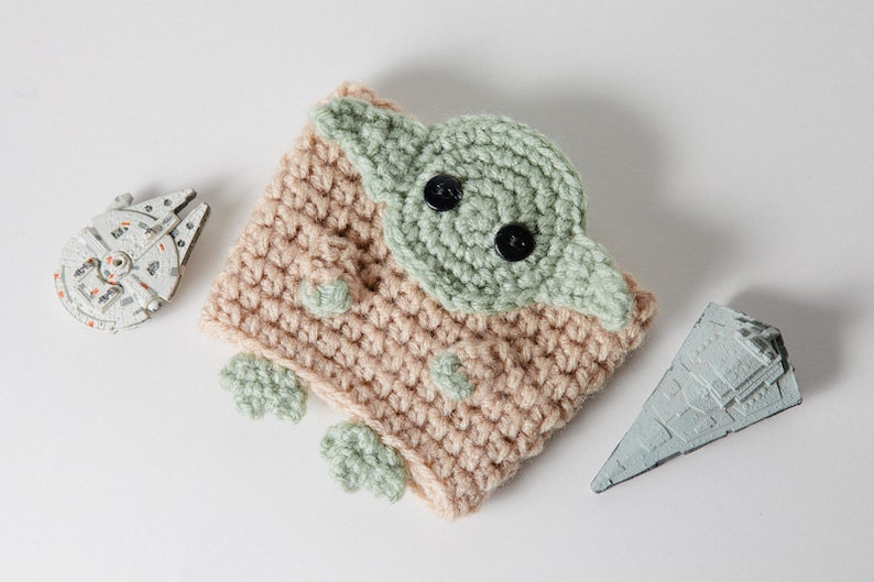 May include: A handmade crochet cup cozy featuring a green and tan design resembling a character from a popular science fiction franchise. The cozy has black button eyes and green accents. Toy models of a spaceship and a star destroyer are also present.