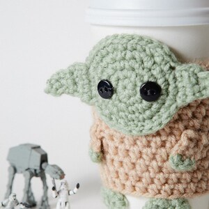 May include: A white coffee cup with a crocheted green and tan Grogu cozy. The cozy features large black button eyes and large ears. Small Star Wars figurines are in the background.