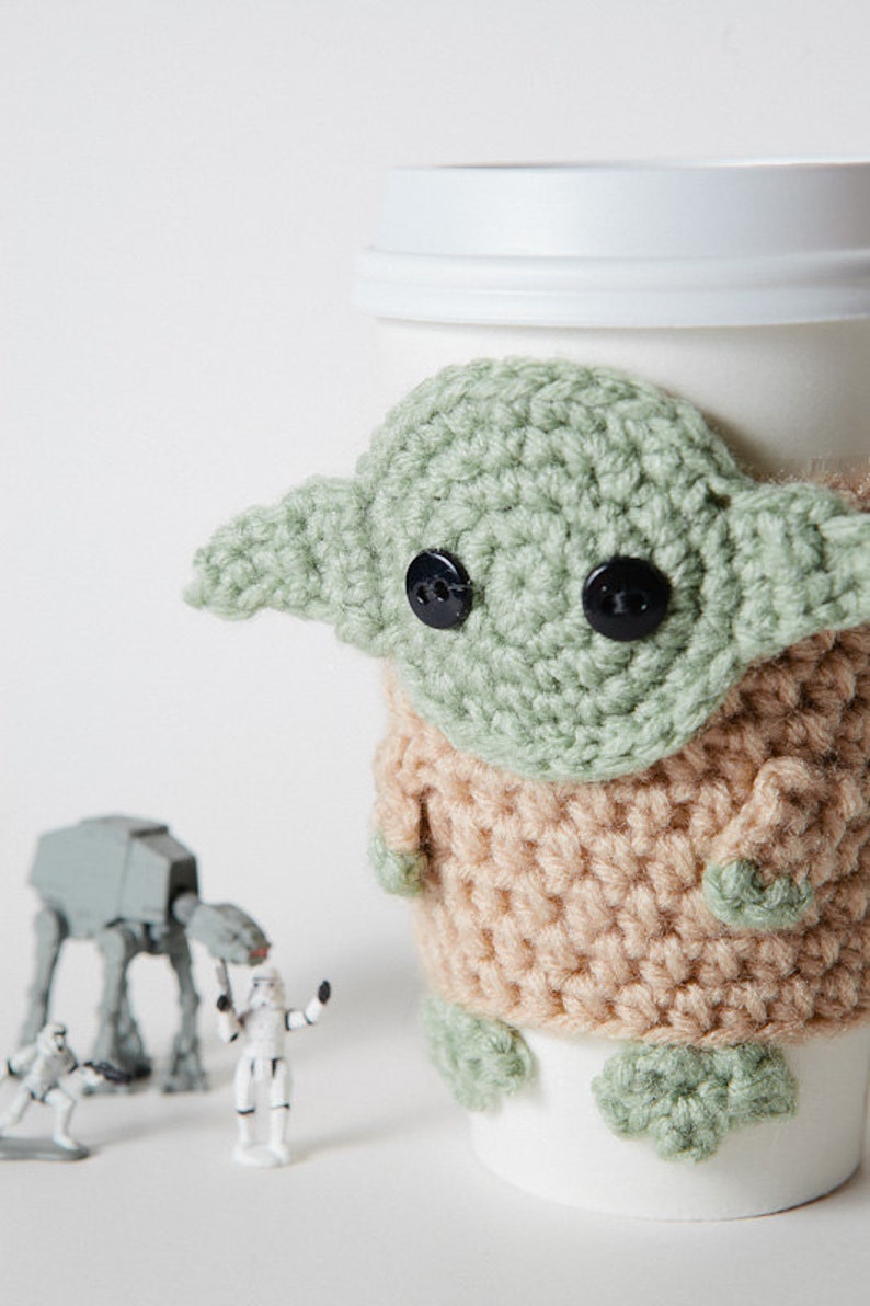 May include: A white coffee cup with a crocheted green and tan Grogu cozy. The cozy features large black button eyes and large ears. Small Star Wars figurines are in the background.