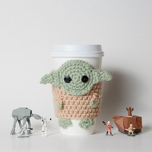 May include: A white paper coffee cup with a crocheted cozy featuring a green and tan design resembling a character from a popular science fiction franchise. Small figurines of characters and vehicles from the same franchise are arranged around the cup.