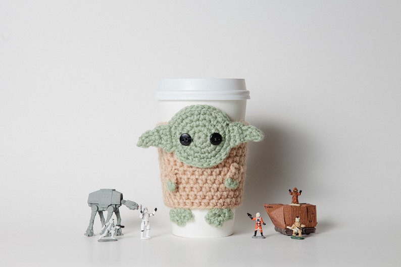 May include: A white paper coffee cup with a crocheted cozy featuring a green and tan design resembling a character from a popular science fiction franchise. Small figurines of characters and vehicles from the same franchise are arranged around the cup.