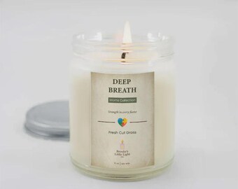 May include: A lit, glass jar candle with a label that reads "DEEP BREATH" from the "Moms Collection." The label also includes the text "Strength in every flame" and "Fresh Cut Grass." The candle is 255 g.