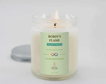 May include: A lit, clear glass jar candle with a silver lid. The candle has a label that reads "Robin's Flame" and "Campfire Marshmallow." The label also includes the text "Advocacy Collection" and "spark of courage in every light."