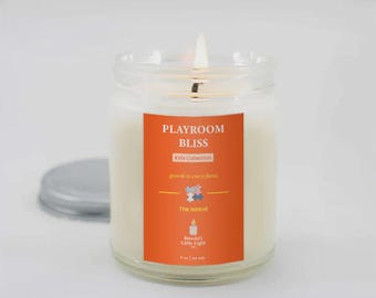 May include: A lit candle in a clear glass jar with a silver lid. The candle has a bright orange label with the words "PLAYROOM BLISS" and "Kids Collection". The label also includes the text "growth in every flame" and "The Island".