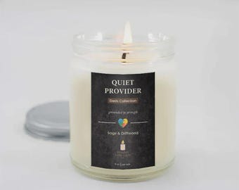 May include: A burning white candle in a clear glass jar with a silver lid. The candle features a black label with the text "QUIET PROVIDER" and "Dads Collection". The label also states the scent "Sage & Driftwood".