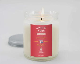 May include: A lit, clear glass jar candle with a red label that reads "A WIN IS A WIN" and "Birthday Cake." The candle is part of the Kids Collection and has a silver lid next to it. The candle is 255 grams.