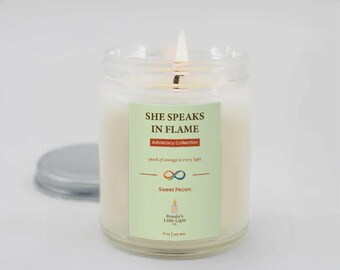 May include: A lit candle in a clear glass jar with a silver lid. The candle has white wax and a label that reads "SHE SPEAKS IN FLAME" and "Sweet Pecan". The candle is part of the "Advocacy Collection".