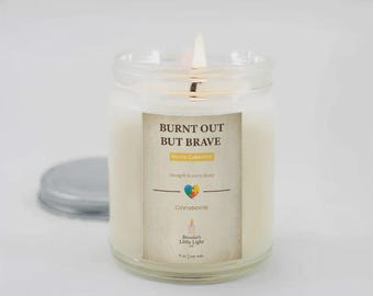 May include: A burning white candle in a clear glass jar with a silver lid. The label says "BURNT OUT BUT BRAVE" with "Moms Collection" and "Strength in every flame". The candle is labelled "Cinnabomb" and "Brooke's Little Light".
