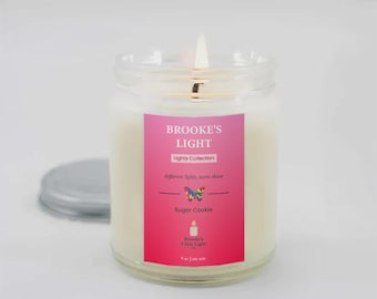 May include: A lit jar candle with a pink label that reads "Brooke's Light" and "Sugar Cookie". The candle is in a clear glass jar with a silver lid. The candle is part of the "Lights Collection".