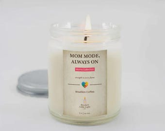 May include: A lit candle in a clear glass jar with a silver lid. The label reads "MOM MODE, ALWAYS ON" and "Brazilian Coffee." The candle is part of the "Moms Collection" and has a colorful heart design. The jar contains 255 grams of soy wax.