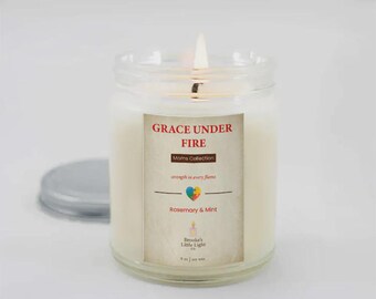 May include: A lit, clear glass jar candle with a white wax interior. The label reads "GRACE UNDER FIRE" and "Rosemary & Mint." The candle is part of the "Moms Collection" and has a silver lid next to it.