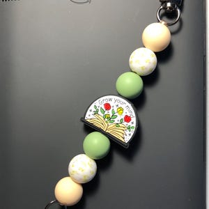May include: A black Amazon Kindle e-reader with a beaded charm. The charm features a black and white graphic with the text "Grow your mind" and is surrounded by cream, green, and white beads. The charm is attached with a silver clasp.