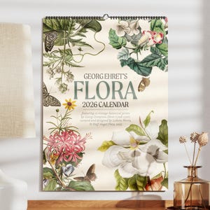 May include: A 2026 calendar entitled "Georg Ehret's Flora" with botanical illustrations of flowers and butterflies. The calendar is displayed next to a white lamp and a decorative vase.