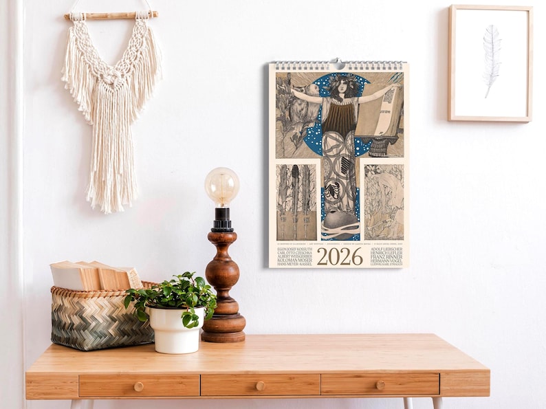 May include: A 2026 wall calendar featuring a central illustration of a figure with outstretched arms against a blue moon, surrounded by intricate line art panels. The calendar includes the year 2026 and various names printed in black text at the bottom.