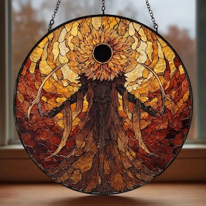 May include: A round stained glass panel featuring a figure with a sunflower-like head and outstretched arms, holding curved blades. The artwork uses warm tones of orange, brown, and yellow, with a black center. The panel is suspended by a chain.