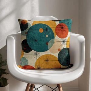 Puede incluir: Un cojín decorativo con un diseño moderno de mediados de siglo. El cojín presenta un patrón de círculos superpuestos en verde azulado, naranja, amarillo y negro, con detalles de estrellas, sobre un fondo beige claro. El cojín está colocado en una silla blanca.