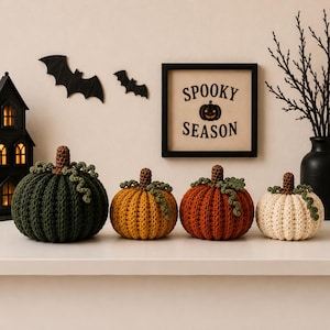 Peut inclure: Quatre citrouilles au crochet en vert foncé, jaune moutarde, orange brûlé et crème, chacune avec une tige brune et des vrilles vertes. Derrière se trouvent une lanterne maison noire, deux chauves-souris noires et un cadre SPOOKY SEASON.