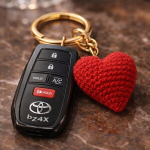 Peut inclure: Un porte-clés de voiture noir avec un porte-clés doré et un charm en forme de cœur rouge crocheté. Le porte-clés comporte des boutons pour verrouiller, déverrouiller et la climatisation, avec le logo Toyota et "bz4X" imprimé.