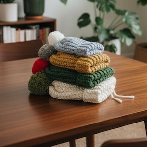 May include: A stack of colorful knit beanies with pom-poms. The beanies are in various colors, including blue, mustard yellow, green, and cream. The beanies are stacked on a dark wooden table.