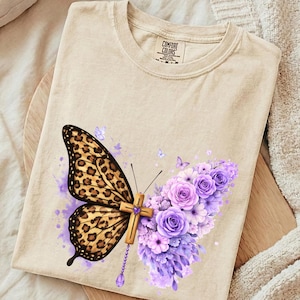 Könnte beinhalten: Beiges T-Shirt mit Schmetterlingsdesign. Ein Flügel hat einen Leopardenmusterdruck, der andere ein Kreuz. Lila Blumen und ein Juwel vervollständigen das Design. Das T-Shirt hat das Markenetikett "Comfort Colors".