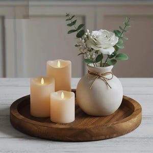 Może przedstawiać: Dekoracyjna aranżacja z trzema kremowymi świecami LED o różnych wysokościach z migoczącym światłem, białym wazonem z kompozycją kwiatową i drewnianą tacą. Wazon jest związany sznurkiem, a świece ułożone są na okrągłej drewnianej tacy.