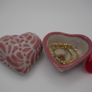 Pode incluir: Duas jarras de cer&acirc;mica em forma de cora&ccedil;&atilde;o, uma com um design floral rosa e branco e a outra com joias de ouro. Uma p&eacute;tala de rosa vermelha descansa perto. Os pratos est&atilde;o em um fundo branco.
