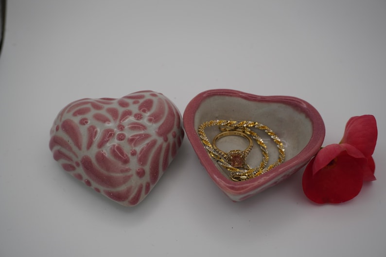 Pode incluir: Duas jarras de cer&acirc;mica em forma de cora&ccedil;&atilde;o, uma com um design floral rosa e branco e a outra com joias de ouro. Uma p&eacute;tala de rosa vermelha descansa perto. Os pratos est&atilde;o em um fundo branco.