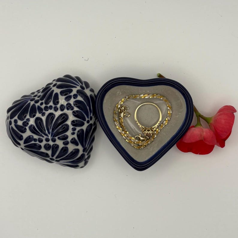 Pode incluir: Uma caixa de joias em forma de cora&ccedil;&atilde;o com um design floral azul e branco ao lado de uma caixa em forma de cora&ccedil;&atilde;o aberta com um anel e uma pulseira de ouro. Uma flor vermelha est&aacute; ao lado da caixa aberta.