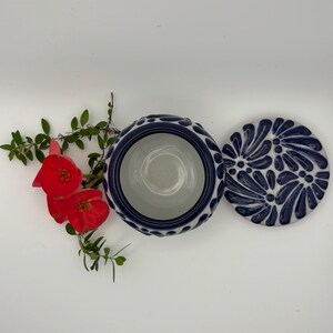 May include: A ceramic bowl with a lid, featuring a blue and white floral design. The bowl's rim has a textured pattern. Red flowers and green leaves are arranged near the bowl.