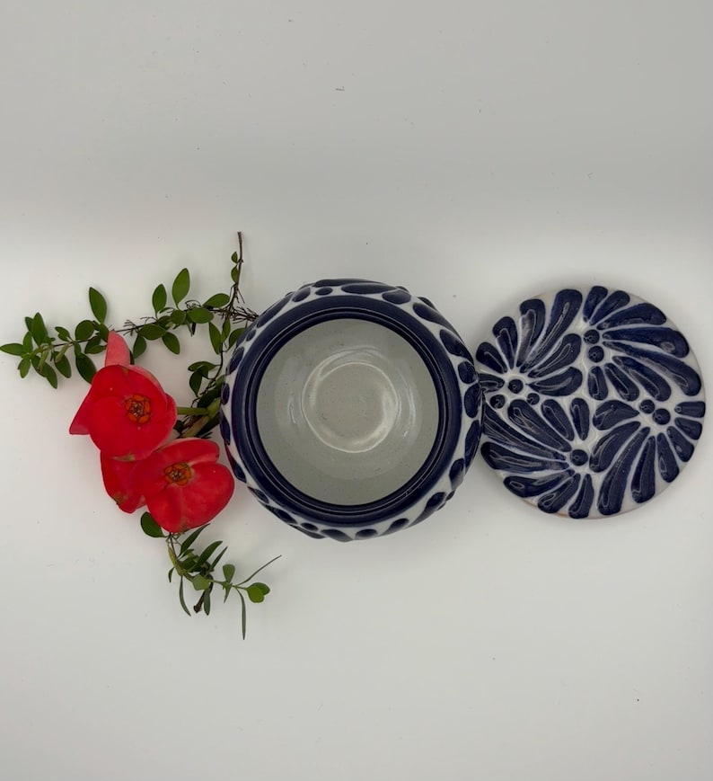 May include: A ceramic bowl with a lid, featuring a blue and white floral design. The bowl's rim has a textured pattern. Red flowers and green leaves are arranged near the bowl.