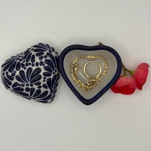 Pode incluir: Uma caixa de joias em forma de cora&ccedil;&atilde;o com uma borda azul e um anel dourado dentro. Ao lado, uma caixa de cer&acirc;mica em forma de cora&ccedil;&atilde;o separada com um design floral azul e branco. Um ramo de flores vermelhas tamb&eacute;m est&aacute; presente.