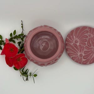 May include: A pink ceramic container with a matching lid, both featuring a textured floral design. The container is open, revealing its interior. Bright red flowers and green leaves are arranged on a white surface, adding a natural contrast to the ceramic pieces.