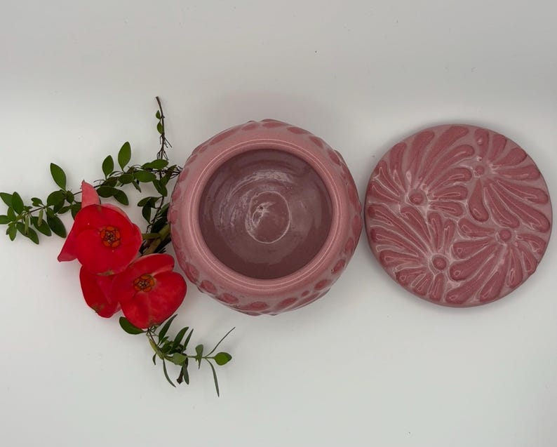 May include: A pink ceramic container with a matching lid, both featuring a textured floral design. The container is open, revealing its interior. Bright red flowers and green leaves are arranged on a white surface, adding a natural contrast to the ceramic pieces.