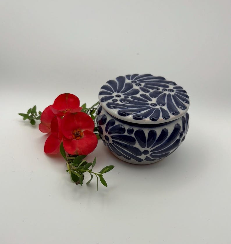 May include: A small, round ceramic jar with a lid, decorated with a navy blue floral pattern. The jar is white with a navy blue floral design. Red flowers and green leaves are next to the jar.
