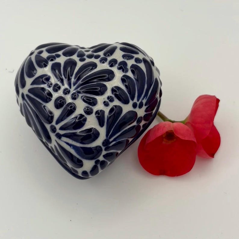 Pode incluir: Um objeto decorativo em forma de cora&ccedil;&atilde;o de cer&acirc;mica com base branca e um padr&atilde;o floral azul escuro. Uma flor vermelha com um caule verde est&aacute; ao lado do cora&ccedil;&atilde;o. O cora&ccedil;&atilde;o tem aproximadamente 7,6 cm de largura.