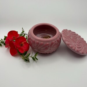May include: A pink ceramic jewelry box with a lid, featuring a floral design. The box is open, revealing a glimpse of gold-colored jewelry inside. Red flowers and green leaves are arranged beside the box, creating a decorative display.