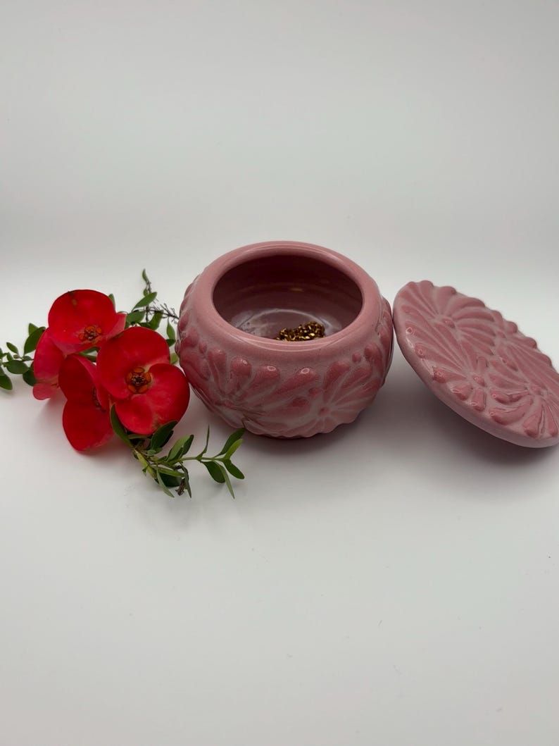 May include: A pink ceramic jewelry box with a lid, featuring a floral design. The box is open, revealing a glimpse of gold-colored jewelry inside. Red flowers and green leaves are arranged beside the box, creating a decorative display.