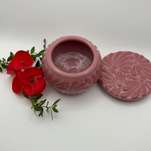 May include: A pink ceramic jar with a matching lid, both adorned with a floral pattern. The jar is open, revealing its interior. Red flowers and green leaves are placed near the jar, adding a touch of color and contrast to the composition.