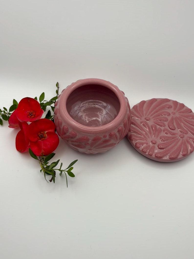 May include: A pink ceramic jar with a matching lid, both adorned with a floral pattern. The jar is open, revealing its interior. Red flowers and green leaves are placed near the jar, adding a touch of color and contrast to the composition.
